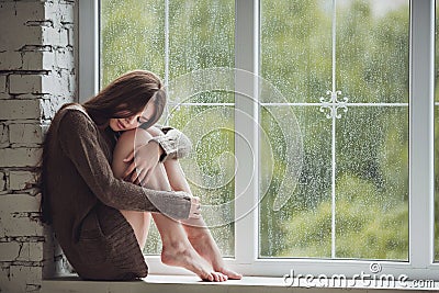 Beautiful young woman sitting alone close to window with rain drops. and sad girl. Concept of loneliness Stock Photo