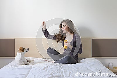 beautiful young woman sitting on bed with her cute small dog besides. Home, indoors and lifestyle. She is drinking orange juice. Stock Photo
