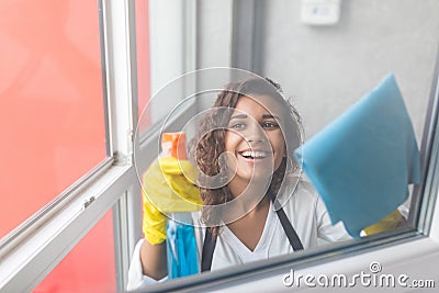 Beautiful young woman is using a duster and a spray, looking at camera and smiling while cleaning windows in the house Stock Photo