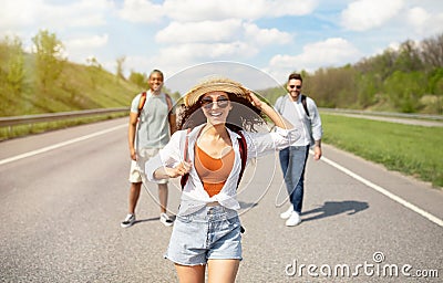 Beautiful young woman walking along highway with her male friends, having summertime journey, backpacking outdoors Stock Photo