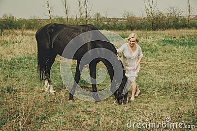 A beautiful young woman in a white chiffon dress with a short blonde haircut strokes and plays with a black horse in a meadow Stock Photo