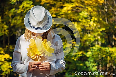 Beautiful young woman in white clothes and hat tilted her head to the bouquet of yellow leaves in her hands. Elegant blonde, face Stock Photo