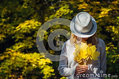 Beautiful young woman in white clothes and hat tilted her head to the bouquet of yellow leaves in her hands. Elegant blonde, face Stock Photo