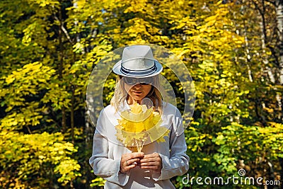 Beautiful young woman in white clothes and hat tilted her head to the bouquet of yellow leaves in her hands. Elegant blonde, face Stock Photo