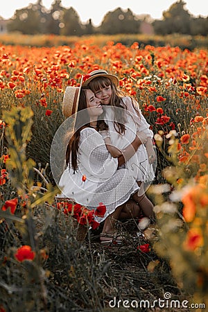 Beautiful young woman with girl in field with poppies, mother and daughter in white dresses and straw hats in evening at sunset, Stock Photo