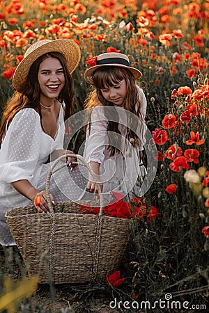 Beautiful young woman with girl in field with poppies, mother and daughter in white dresses and straw hats in evening at sunset, Stock Photo