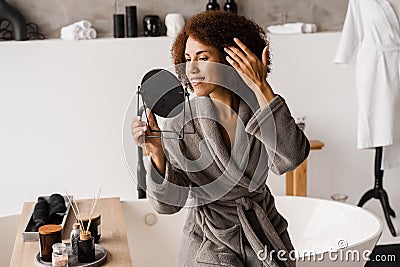 Beauty morning routine of african girl in bathroom. Attractive african american woman in bathrobe is looking in the Stock Photo