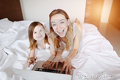 Beauty mother hugs little daughter with laptop laying at white bed with sunshine Stock Photo