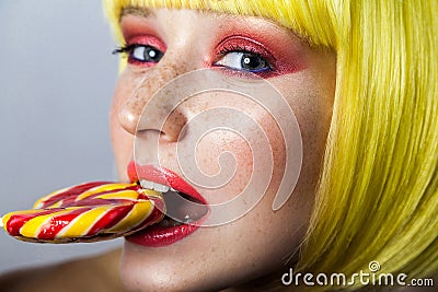 Beauty portrait of cute young female model with freckles, red makeup and yellow wig, holding and bitting her colorful candy stick Stock Photo