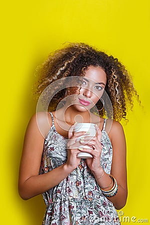 Beauty portrait of young african american woman with afro hairstyle. Girl posing with coffee on yellow background, looking at came Stock Photo