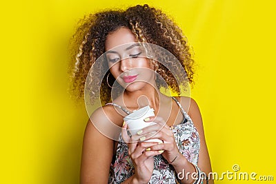 Beauty portrait of young african american woman with afro hairstyle. Girl posing with coffee on yellow background, looking at came Stock Photo