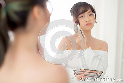 Beauty portrait young asian woman smiling with face looking mirror applying makeup with brush cheek in the bedroom. Stock Photo