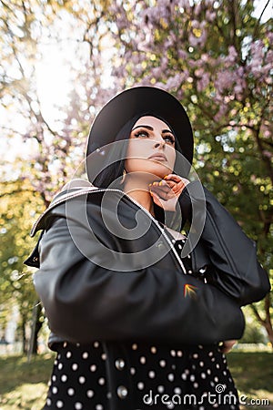 Beauty sunny portrait chic woman in black stylish dress in fashionable hat in vintage leather jacket in park on sunny bright day. Stock Photo