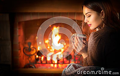 Beauty young Christmas woman sitting near fireplace in dark room at home and drinking hot beverage from a big cup Stock Photo