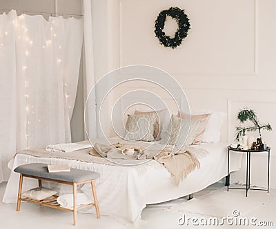 Bed with beige knitted plaid and a cup. Interior of stylish cozy bedroom with wreath on the wall. Christmas, New year home decor Stock Photo