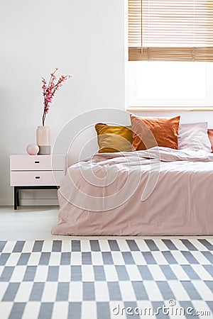 Bed with pink sheets and orange cushions standing in white bedroom interior with carpet, window with wooden blinds and bedside ta Stock Photo
