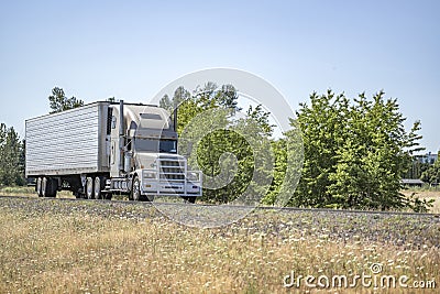 Beige big rig bonnet classic semi truck with grille guard transporting cargo in refrigerator semi trailer driving on the narrow Stock Photo