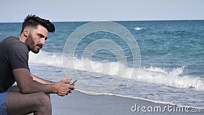 Bemannen Sie beschäftigtes mit seinem Telefon, das an den Strand-Flusssteinen sitzt stock video footage Bemannen Sie beschäftigtes mit seinem Telefon, das an den Strand-Flusssteinen sitzt stock video footage