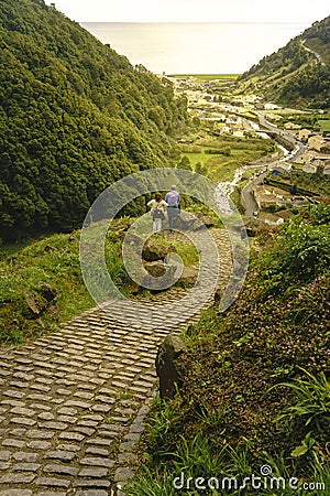 Bending yellow stone pavement pathway descending to the river. Adult couple standing and looking for the sun rays over a tiny vill Editorial Stock Photo