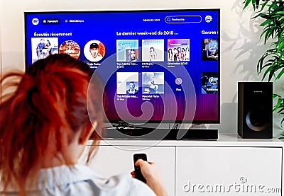 Benon, France - December 30, 2018: Redhead woman sitting in her living room and holding a television remote control facing a Editorial Stock Photo