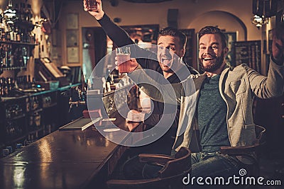 Best friends having fun watching a football game on TV and drinking draft beer at bar counter in pub. Stock Photo