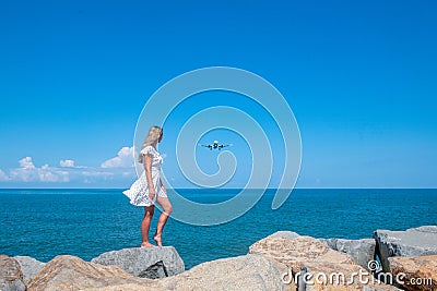 Beyond the Shore: Girl in White Dress, Stones, Sea, and an Airborne Plane Stock Photo