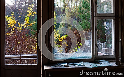 The big broken window in the abandoned building with autumn colorful trees outside it. Stock Photo