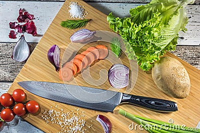 Big chef knife with healthy food - vegetables, onion, salad, potato placed on a cutting board with wood background top view Stock Photo