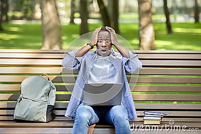 Big failure. Young black guy holding his head in panic near laptop computer at park Stock Photo