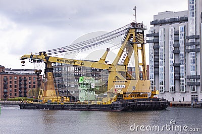 A big heavy yellow crane sailing on a barge through an urban harbor with modern buildings in the background. Editorial Stock Photo