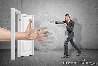 Big male open hand appearing through white doorway and businessman making kicking gesture on grey wall background Stock Photo