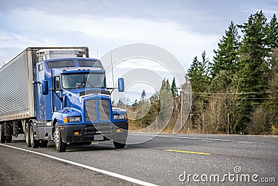 Big rig blue classic powerful semi truck transporting frozen cargo in refrigerated semi trailer running on the road with trees on Stock Photo