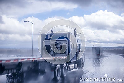 Big rig blue classic semi truck with flat bed semi trailer moving on the wet raining road behind another semi truck with rain dust Stock Photo