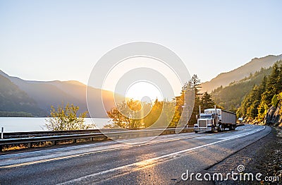 Big rig classic powerful semi truck transporting cargo in covered long bulk semi trailer running on the road along the river with Stock Photo