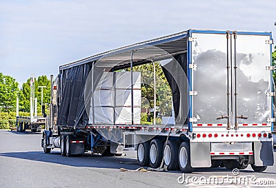 Big rig semi truck with open covered semi trailer unloading commercial cargo on warehouse parking lot Stock Photo