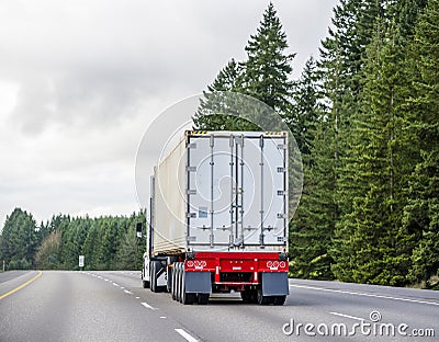 Big rig semi truck transporting cargo in long container mounted on a flat bed semi trailer running on the wide road with green Stock Photo