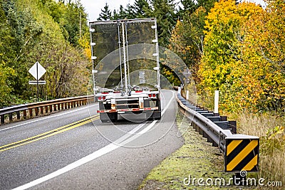 Big rig semi truck transporting cargo in semi trailer with mirrored metal tailgate running on the winding autumn road with trees Stock Photo