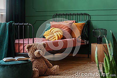 Big teddy bear and an open book on a green, velvet pouf in front of a metal frame bed in child`s bedroom interior. Real Stock Photo