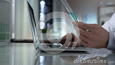 Biochemist holding tube with turquoise liquid, typing test results on laptop Stock Photo