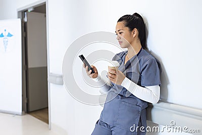 Biracial female doctor wearing scrubs using smartphone, holding takeaway coffee, copy space Stock Photo