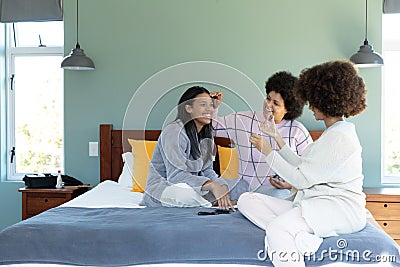 Biracial female friends applying make-up and painting fingernails of happy young woman on bed Stock Photo