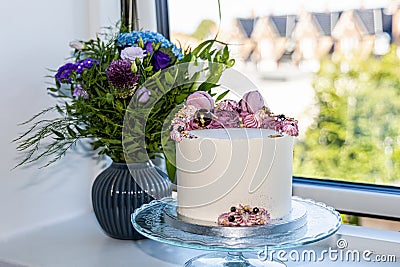 Birthdag cake decorated with pink flowers and a bouguet of flowers in a vase on the windowsill. Stock Photo