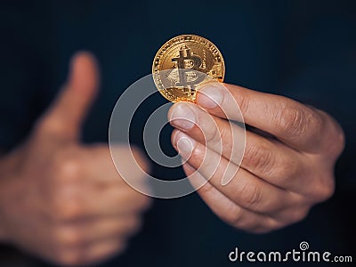 Bit coin on one hand, other hands in thumbs up gesture. Selective focus. Dark blue background. Money and finance concept. Stock Photo