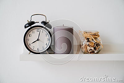 Black alarm clock, jar with dried flowers and perfume and gray candle on shelf. Stock Photo