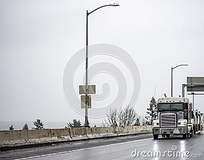 Black big rig classic semi truck with turned on headlights transporting cargo on flat bed semi trailer running on the slippery wet Stock Photo