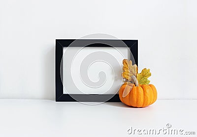 Black blank wooden frame mockup with an orange pumpkin and golden oak leaves lying on the white table. Poster product Stock Photo