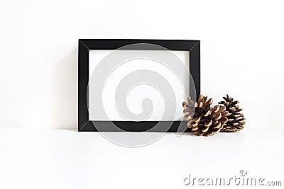 Black blank wooden frame mockup with pine cones lying on the white table. Poster product design. Styled stock feminine Stock Photo