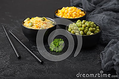 Black bowls with boiled organic basmati vegetable rice, yellow corn and peas with black chopsticks on black stone background with Stock Photo