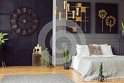 Black clock, golden chandelier, paintings and white bed in an elegant bedroom interior. Real photo Stock Photo