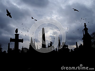 Crows flying and perched on old gothic style gravestone in silhouette with tall memorials and crosses against an overcast cloudy Stock Photo